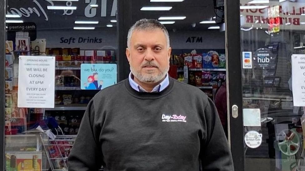 Mohammed is wearing an ironed black sweatshirt with the “Day-Today” logo over the breast pocket. He’s standing in front of his shop. He’s an older middle aged man with short black hair and a greying beard.