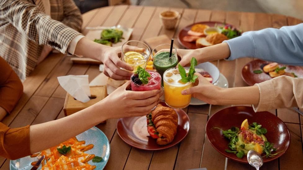Close up of four women putting their drinks together in a 'cheers' motion. They are sat around a wooden table with plates of food in front of them.