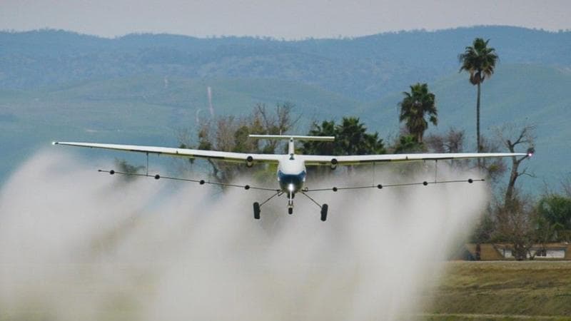 A Pyka crop-duster flies low over a field spraying liquid.