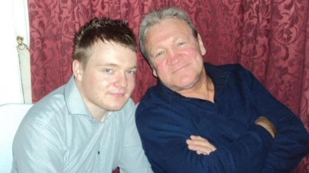 A picture of a father and son. The younger man wears a pale grey shirt and the older man wears a navy shirt with his arms crossed. Both look relaxed and happy.
