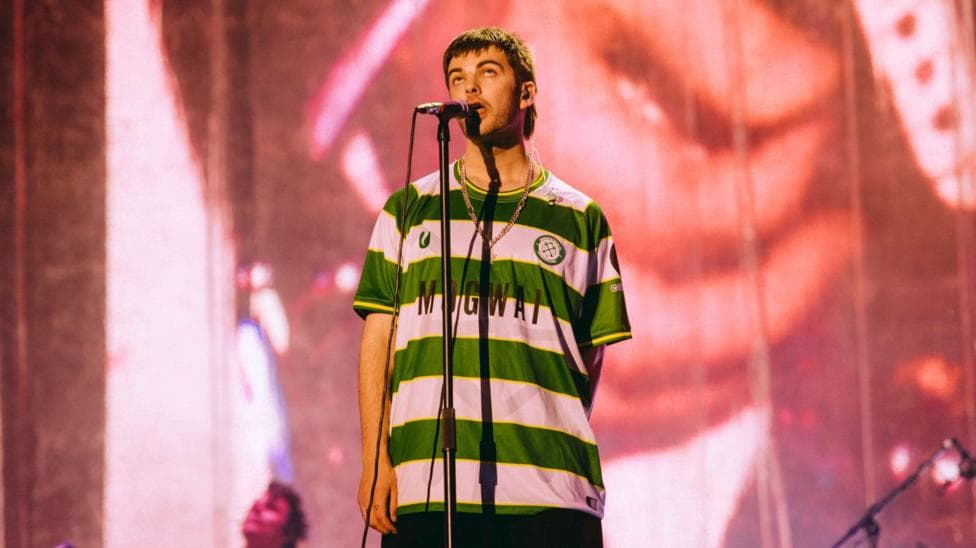 Grian Chatten performing at Reading Festival wearing a Cwmbran Celtic FC away shirt. The football shirt is green and white.
