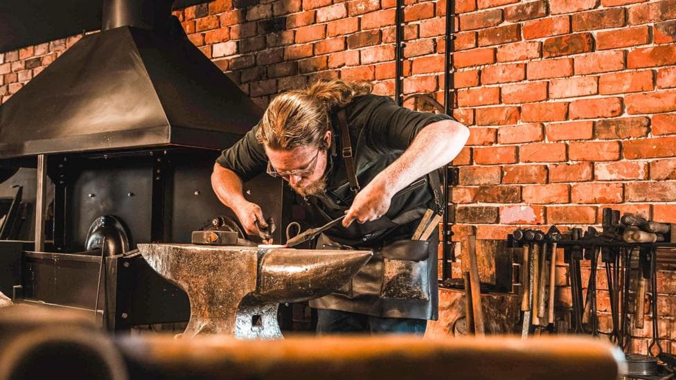 A man with long, red hair tied back leans over an anvil working in detail on metal. Behind him are a brick wall, a set of tools and a black forge.