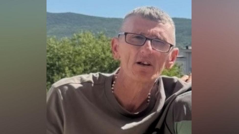 James Bingham. He has short grey hair, wearing glasses, a silver chain and khaki t-shirt. Tress and mountains are behind him.