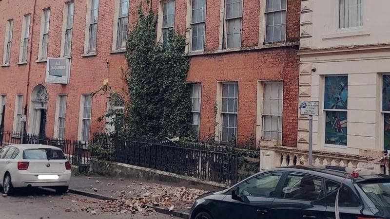 A street photo showing a row of terraced Georgian-style buildings. A pile of bricks lie on the pavement below the building in the middle of the photo. Cars are parked on either side of the bricks.