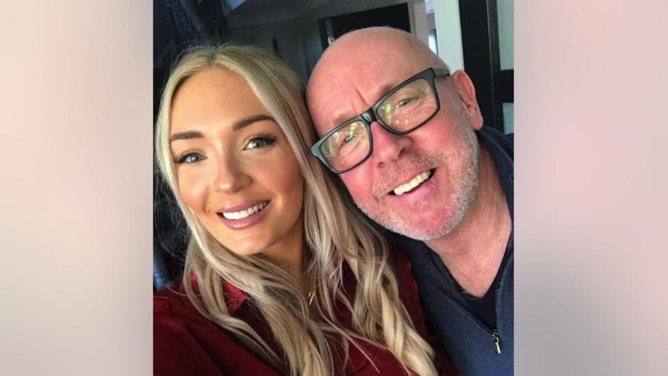 A young woman with long blonde hair smiles and poses with her father, an older man wearing black rimmed glasses.