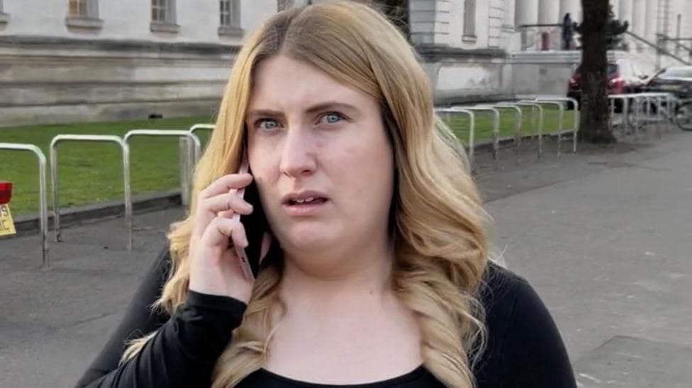 Charlotte Blackwell looks at the camera as she is on the phone. She is standing in front of a court and she has long blonde hair and is wearing a black top.