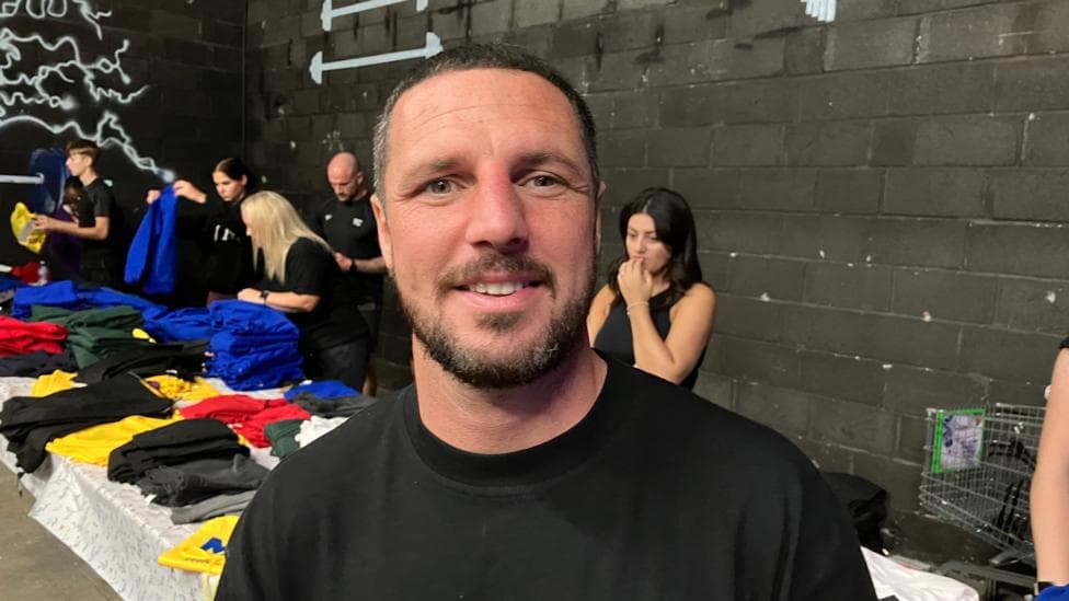 A man with short black hair and a black beard, wearing a black T-shirt. Behind him is a long table with various multi-coloured items of school uniform laid out. Five people are behind the table, handing out the items of clothing.
