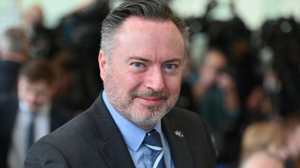A smiling man with greying dark hair and beard is wearing a grey suit jacket, blue shirt and blue striped tie