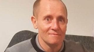 Head and shoulders shot of a smiling bald man sitting on a dark grey couch against a dark grey wall.