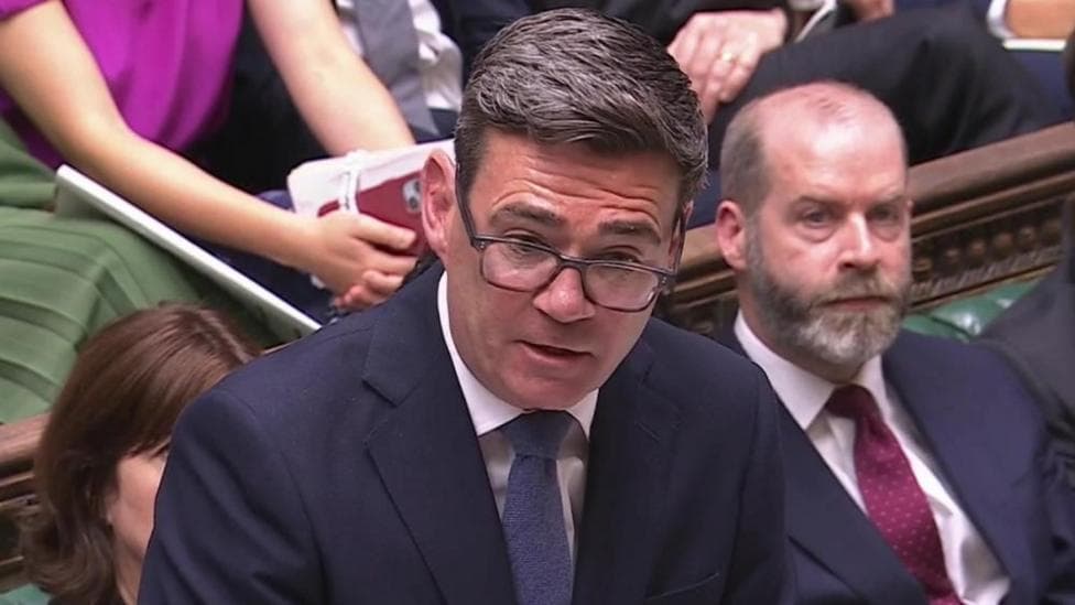 Andy Burnham is wearing a dark coloured suit jacket, white shirt, blue tie and glasses. He has short, black hair. He is stood up and is speaking. Behind him are MPs sat on the green benches of the House of Commons.