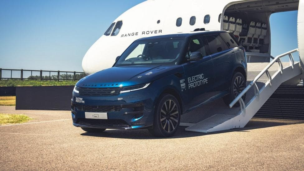 A blue Range Rover Electric prototype drives down a metal ramp from the open cargo bay of a large white aircraft. The vehicle carries “Electric Prototype” markings and technical angle indicators on its body. The aircraft is branded “Range Rover” near the windows.