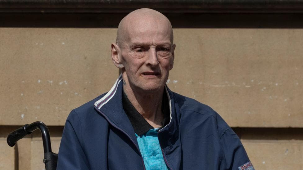 a bald man in a blue tracksuit top and t-shirt is sitting in a wheelchair with a building wall behind him