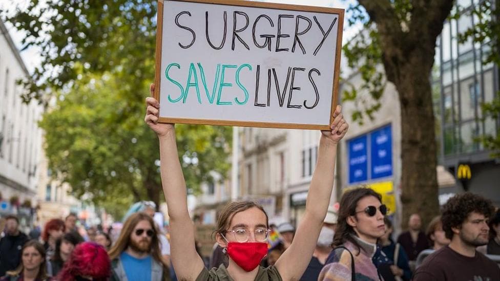 A person holds a sign up that reads "SURGERY SAVES LIVES".