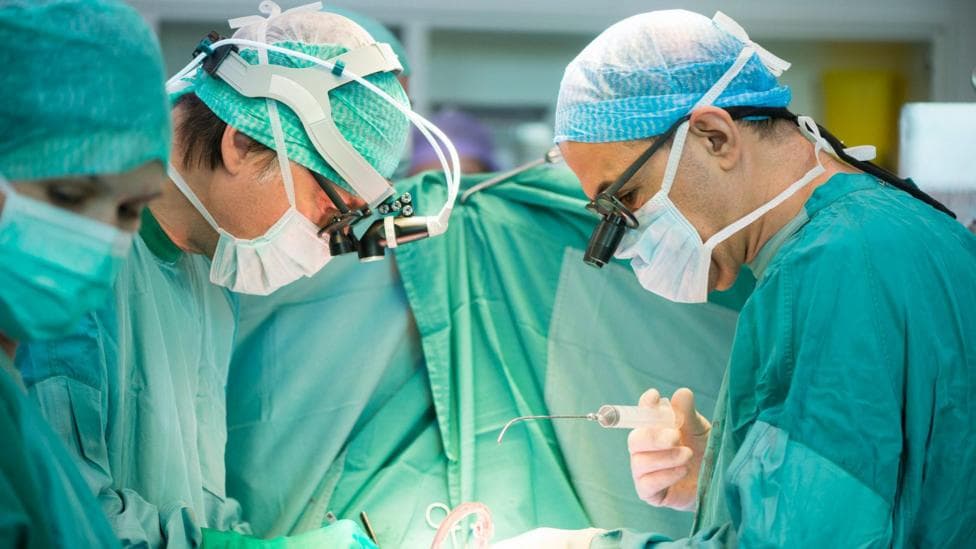 Two surgeons, facing each other, stood over a patient as they operate. They are wearing face masks, goggles and are dressed in green and blue scrubs.