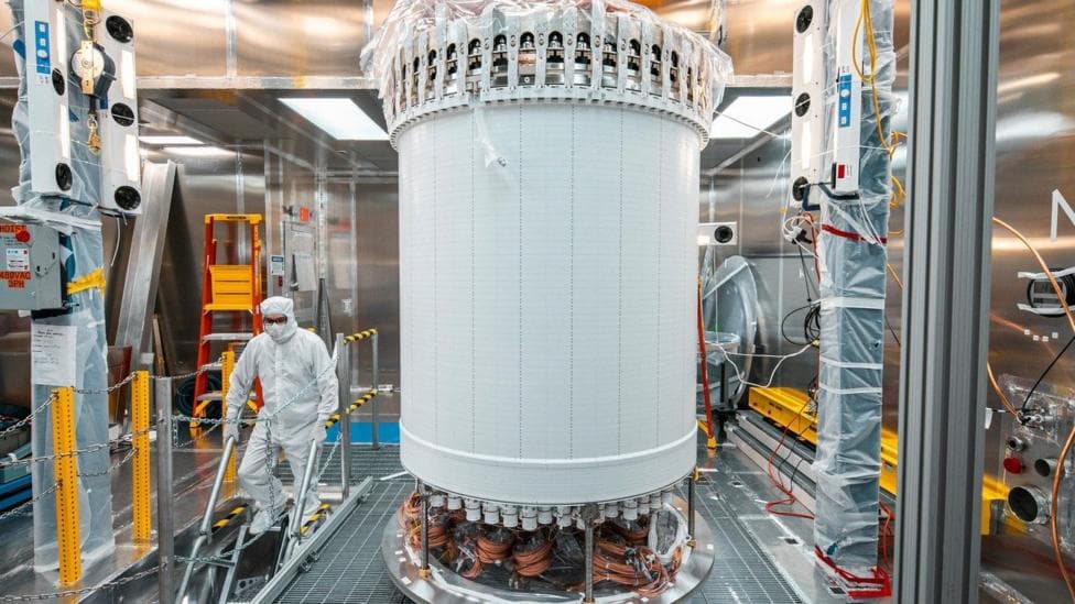 The LUX-ZEPLIN main detector in a laboratory. The LZ is a large white cylindrical machine. A person wearing a white hazmat suit is scene walking down some stairs next to the machine.