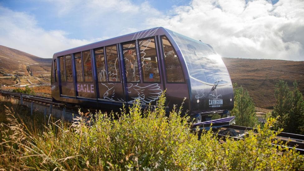 A train carriage-like vehicle travels along a rail on a hillside in the Cairngorms. The vehicel is purple and has a row of windows.