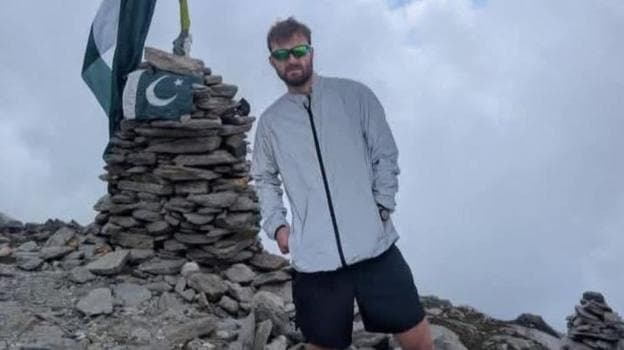 Alexander Harris, a man in black shorts and a grey jacket and wearing sunglasses, is posing in front of a stone formation and Pakistan flags