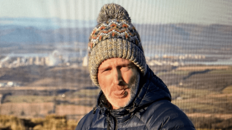 a man in a green wooly bobble hat and black jack is sticking out his tongue as he poses for the camera. There is a hillside/field landscape behind him
