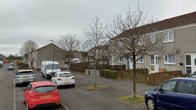 A row of grey terrarced houses, with patches of grass outside them. Cars are parked in front of some of them, with another row of homes in the background