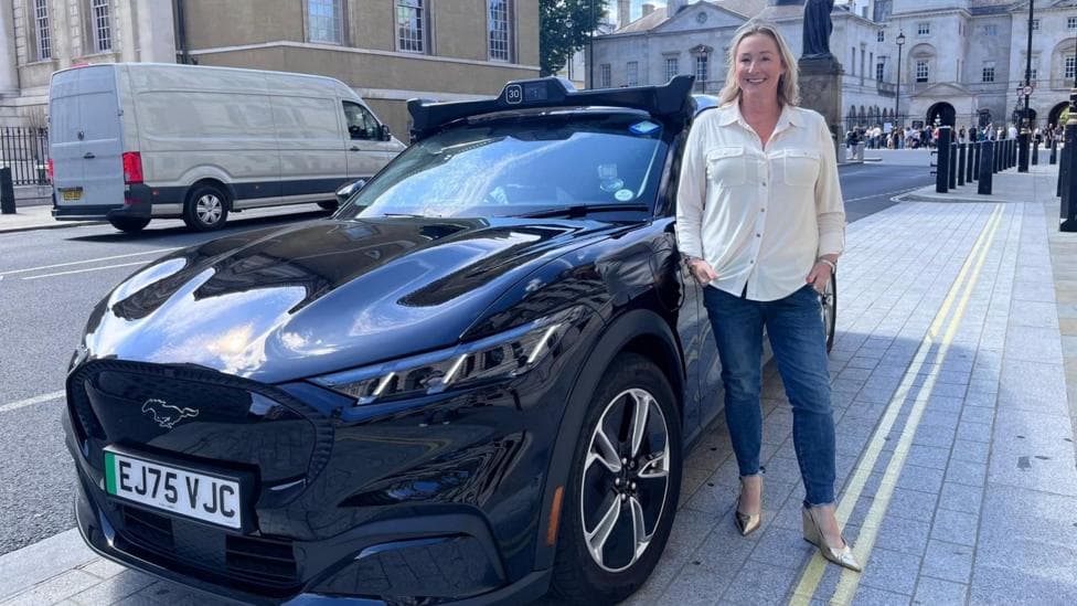Zoe Kleinman in blue jeans and cream blouse standing next to a dark Wayve self-driving robotaxi on a London pavement.