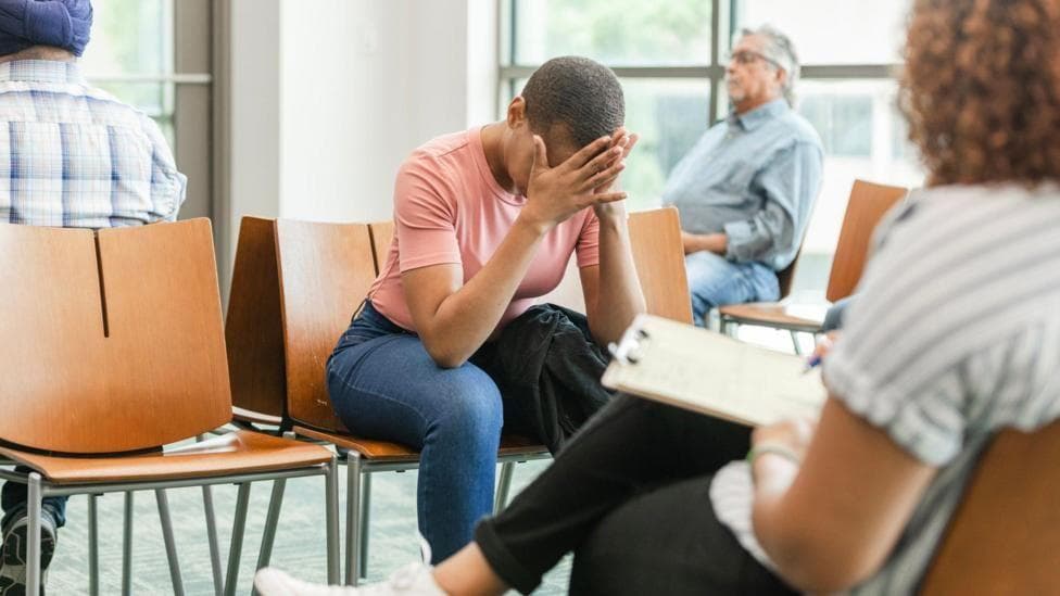 A stock image of people sat in a hospital waiting room. One woman with short dark hair, wearing a pink t shirt, has her head in her hands.
