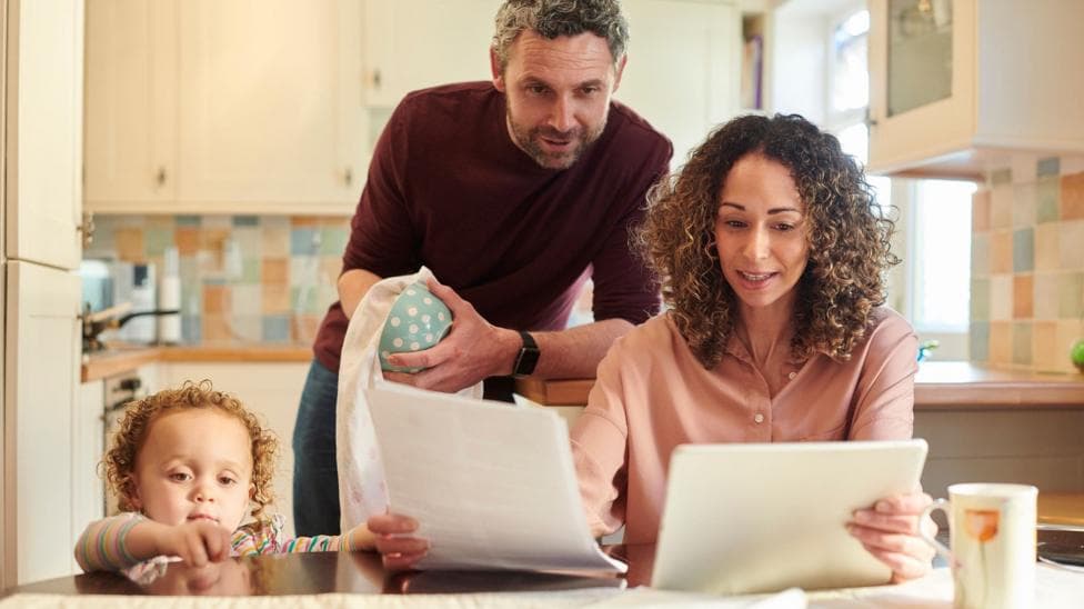 A family of three looking at bills and finances at a kitchen table. From left to right, a child is standing on the floor looking out across the table, a man is looking over a woman's shoulder at A4 sheets of paper she is holding while standing drying a mug, and a woman sits at the table holding the papers and reading. The kitchen behind them in soft focus is mostly pastel-coloured.
