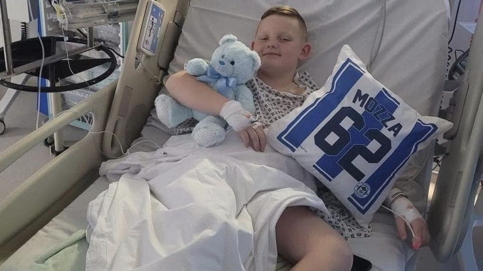 A young boy with short brown hair is smiling while lying in a hospital bed. He is holding a blue teddy bear and a Wigan Athletic cushion with 'Mozza 62' written on it. He has a drip in his right hand and bandages and a medical tube connected to his left hand.