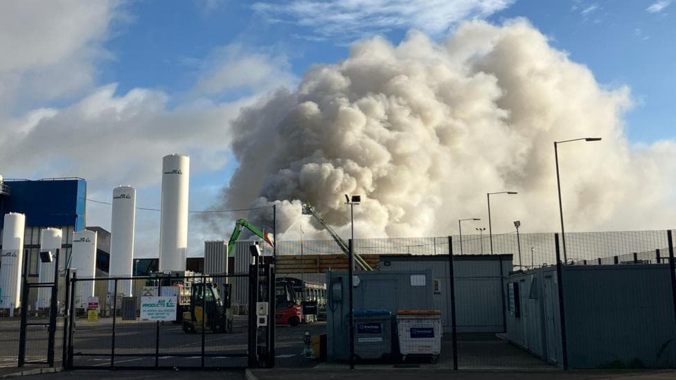 A large white plume of smoke rises into a blue sky from an industrial site.