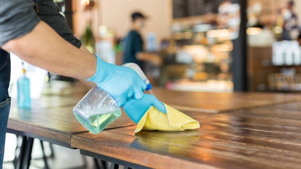 Person wearing blue disposable gloves sprays cleaning solution onto a wooden table and wipes the surface with a yellow cloth inside a café or restaurant, with the background softly blurred.