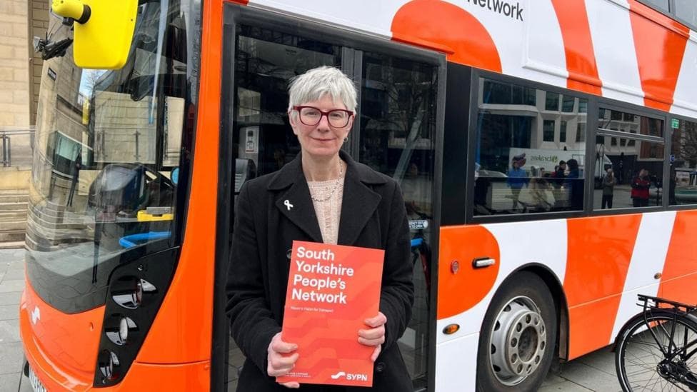 Councillor Leader Fran Belbin is standing in front of an orange and white coloured bus. She has short grey hair and is wearing burgundy-coloured glasses. She is wearing a black jacket and has a cream-coloured top underneath. She is holding a leaflet which is orange and has "South Yorkshire People's Network" written in white