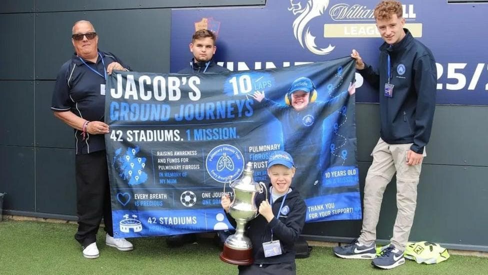 Three men hold a banner which says Jacob's Ground Journeys, 42 stadiums, one mission. A 10-year-old boy wearing a baseball cap holds up a football trophy in the foreground.