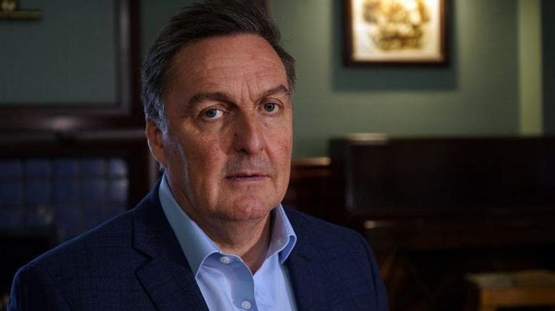 John Morrison is sitting in a dimly lit room. He is looking into the camera. He is wearing a dark blue suit jacket over a light blue shirt.