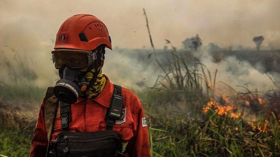 A person in orange firefighting uniform, helmet and gas mask stands amid smoke in front of flaming grass