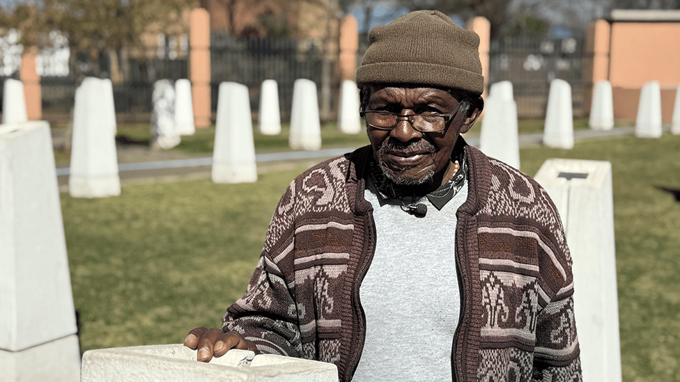 Abraham Mofokeng in a brown patterned cardigan and beanie hat stands by a white monolith. Out of focus, in the background, are other white monoliths - they are part of a memorial.