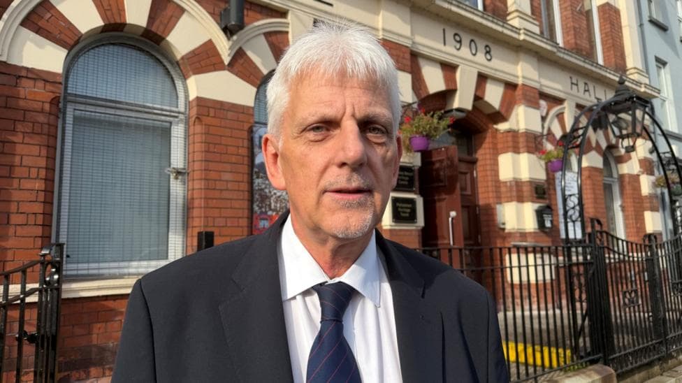 A white haired man looking at the camera. He is wearing a black jacket and white shirt. His tie is navy with red pin stripes.