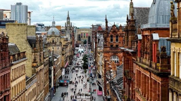 View down Glasgow's Buchanan Street from an elevated position, showing pedestrians along the wide pedestrianised shopping street, flanked by historic sandstone buildings and domed rooftops, with the city centre skyline stretching into the distance under a cloudy sky.