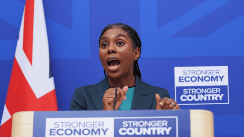 Badenoch is pictured from below the podium where she is giving a speech, at London Scottish House in Westminster, with a royal blue background and a Union flag to her left. She wears her braided hair tied back in a low ponytail, with a navy jacket and a turquoise top.