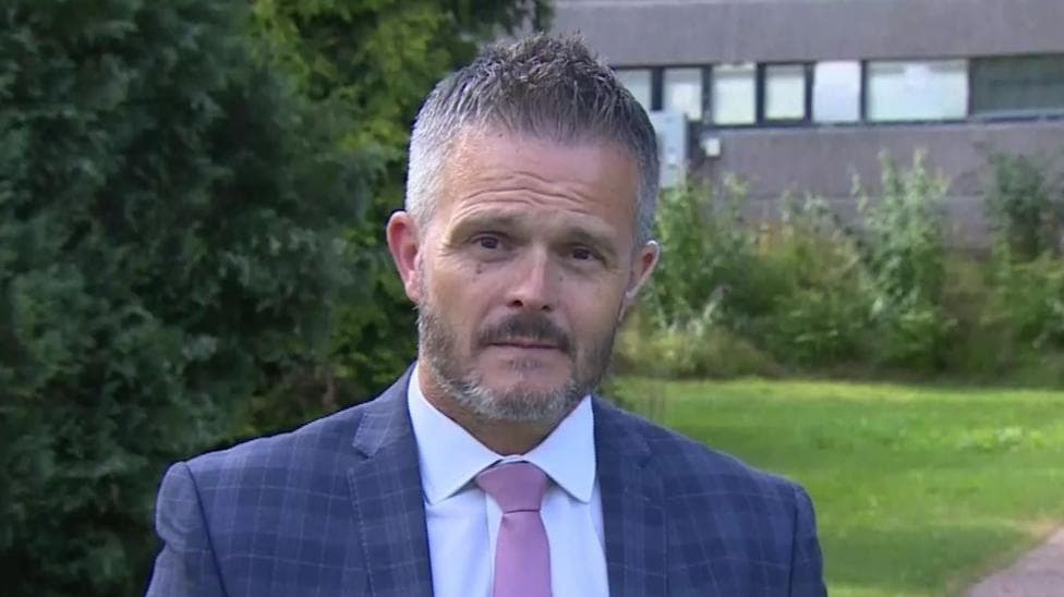 Robbie Butler. He has short grey-ish hair, wearing a blue check blazer, white shirt and light purple tie. Behind him is a dark grey office building and grass.