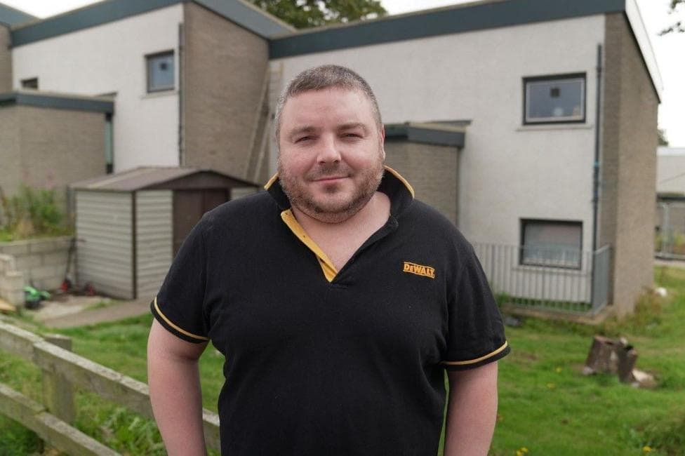 Raymond Smith, a man smiling at the camera, has short brown hair and short facial hair. He is wearing a black short-sleeved shirt and is standing in front of housing.
