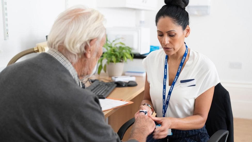 Stock photo shows a GP with an NHS lanyard around their neck in a GP surgery setting, putting a finger monitor onto a patient during a consultation.