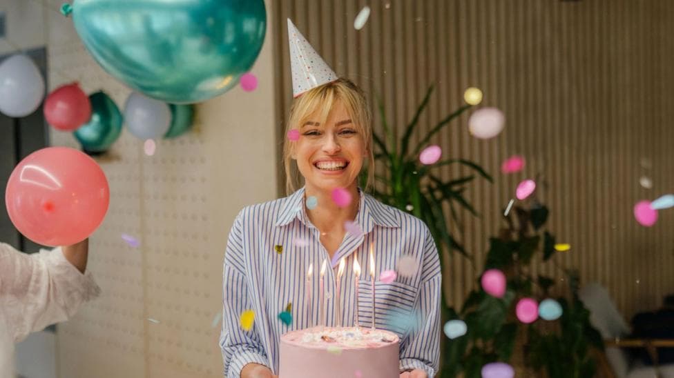 A young woman holding a pink birthday cake with five candles on, wearing a party hat and striped shirt, celebrating her birthday, with balloons seen hanging behind her