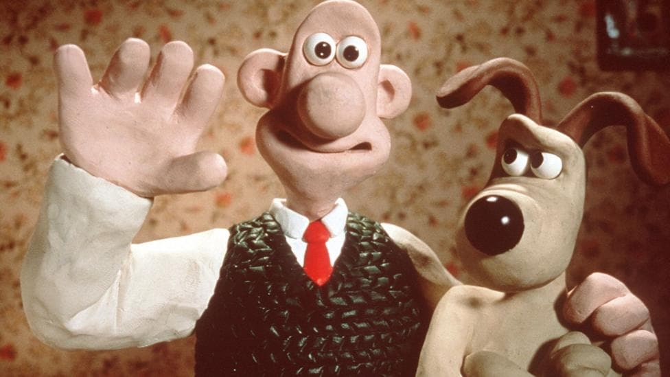 A stopmotion character of Wallace is waving and smiling at the camera with his arm round Gromit. Gromit the dog is giving him some serious side eye