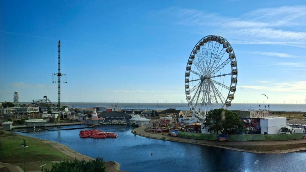 A seaside scene in Skegness with a big wheel and a funfair, the skies are blue