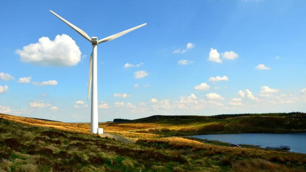 A windturbine sits on a grassy area surrounded by a lake. This sky is blue dotted by clouds