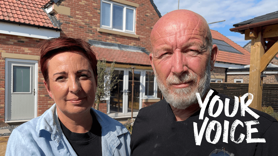 Elissa and Johnnie stand in front of a modern brick house with red‑tiled roofing and solar panels. Elissa wears a light blue shirt, the Johnnie a black shirt with safety glasses tucked into the pocket. The house has white‑framed windows, a light‑coloured door, and a clear blue sky overhead.