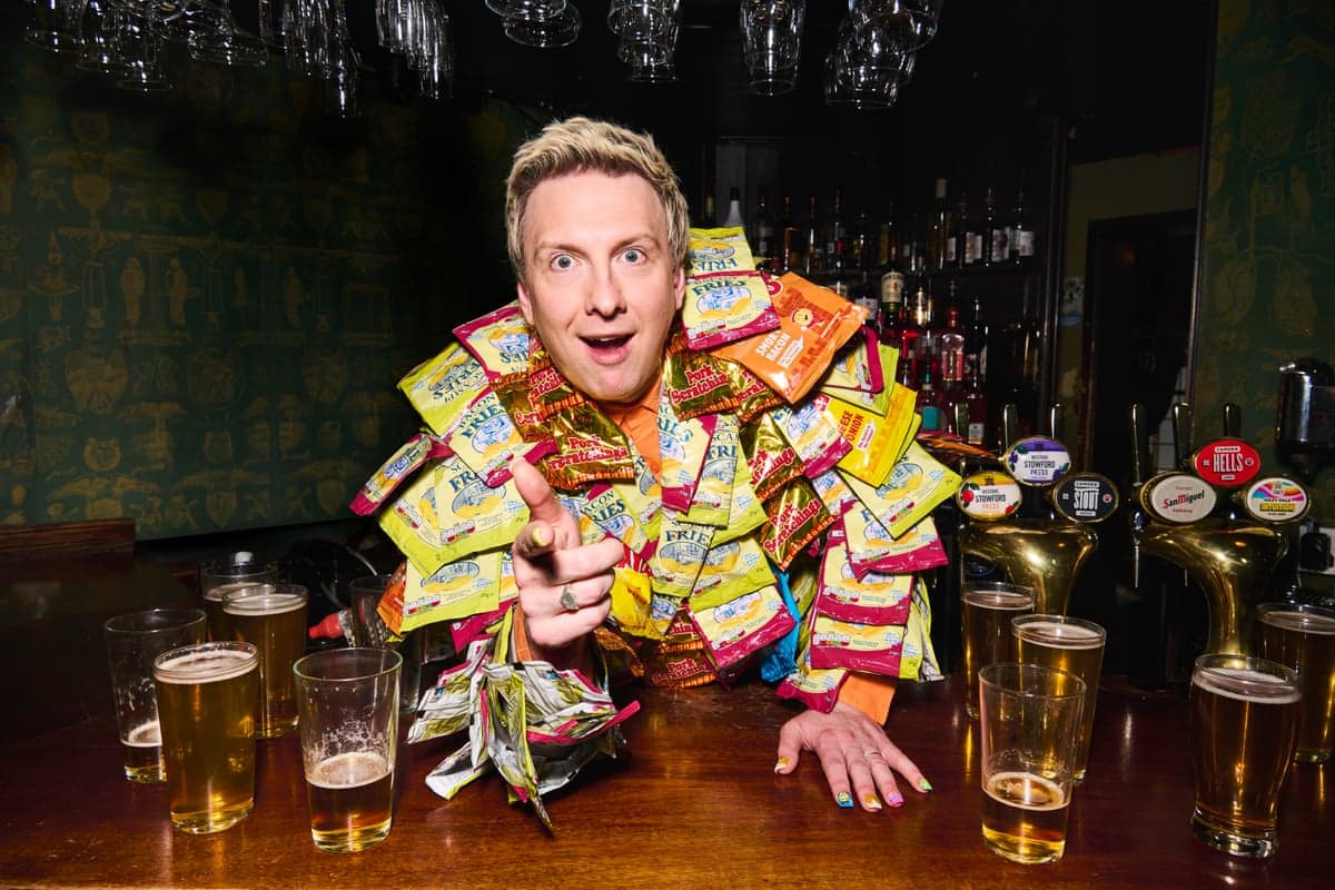 A man with short blond hair covered in packets of nuts, behind a bar, with pints of beer either side of him