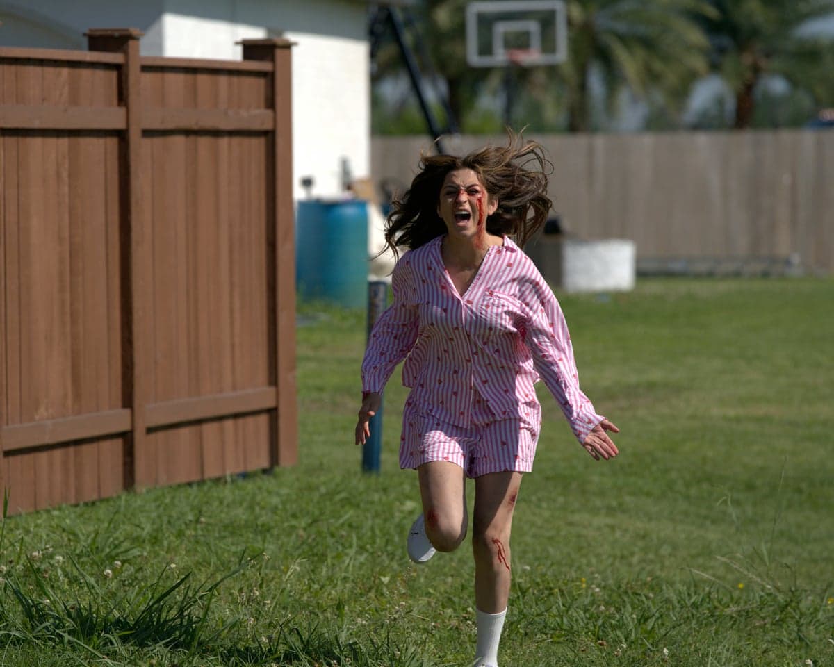 A woman in blood-spattered pajamas runs screaming across a grassy backyard