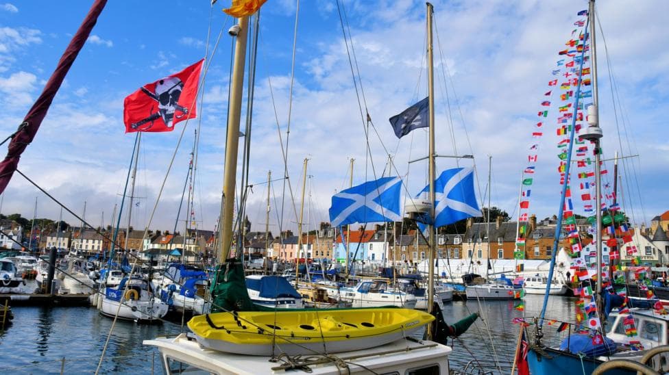 Different coloured fishing boats with flags and bunting moored in Anstruther Harbour on a sunny day. Colourful buildings can be seen in the background.