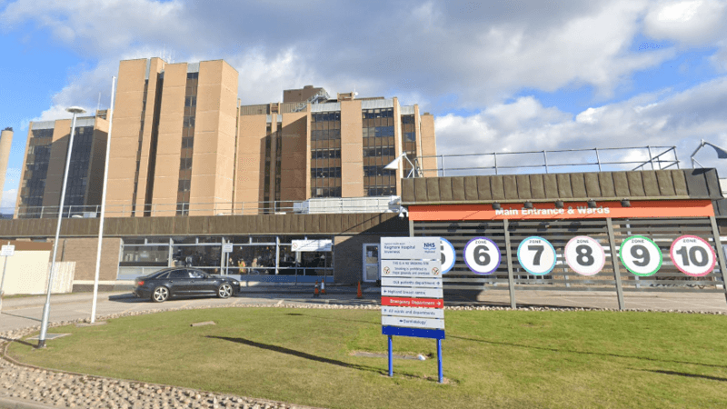 An exterior view of Raigmore Hospital in Inverness. It is a tall, boxy building. At ground level is the entrance with round signs with numbers indicating different zones inside the hospital.