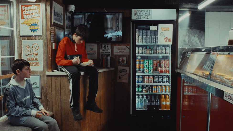Still from a video showing two boys sitting in a chip shop with a fridge full of drinks and a number of posters